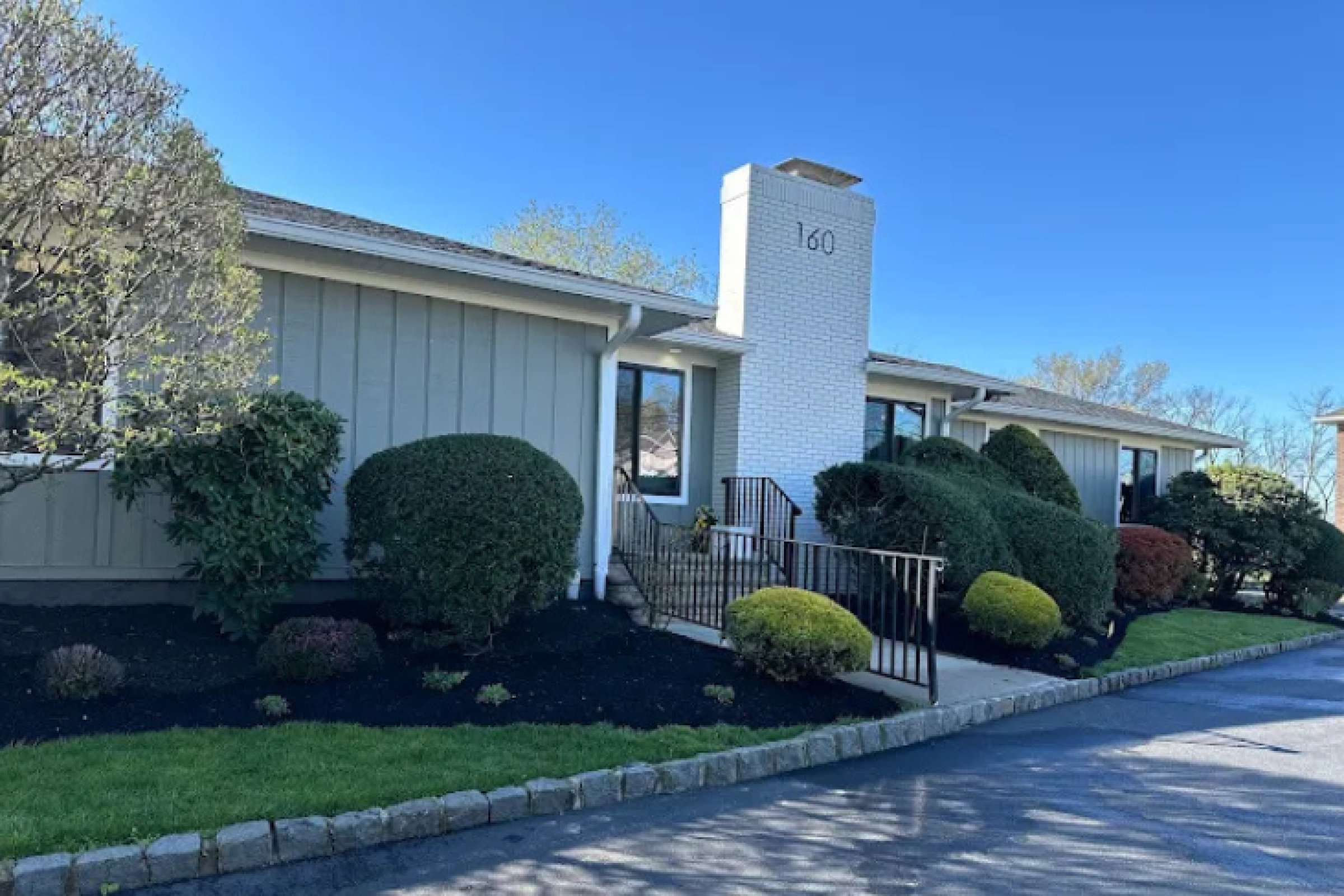 The exterior of a dental clinic located at 160, featuring well-maintained landscaping and a bright blue sky.