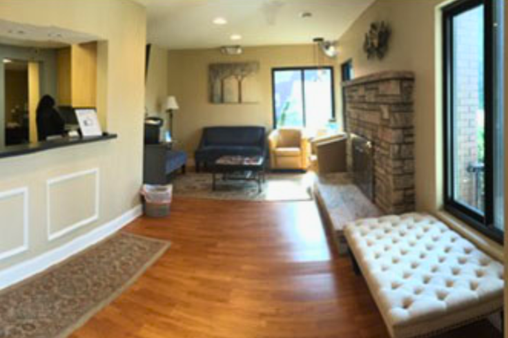 The image depicts a well-lit dental clinic waiting area featuring a reception desk, wood flooring, comfortable seating, and a fireplace.