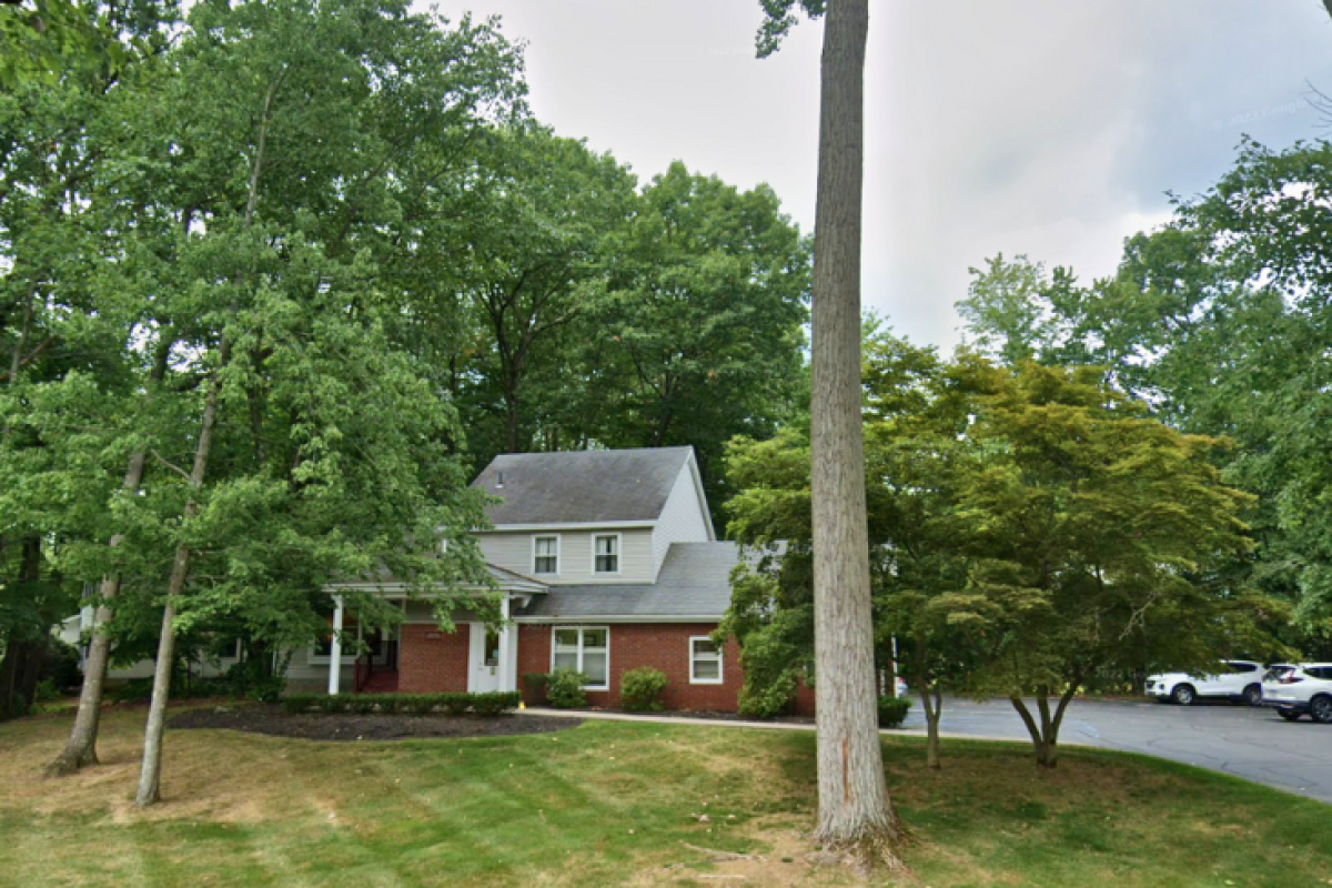A charming brick and white-paneled building surrounded by lush greenery and trees, with a neatly maintained lawn and a few parked cars in the driveway.