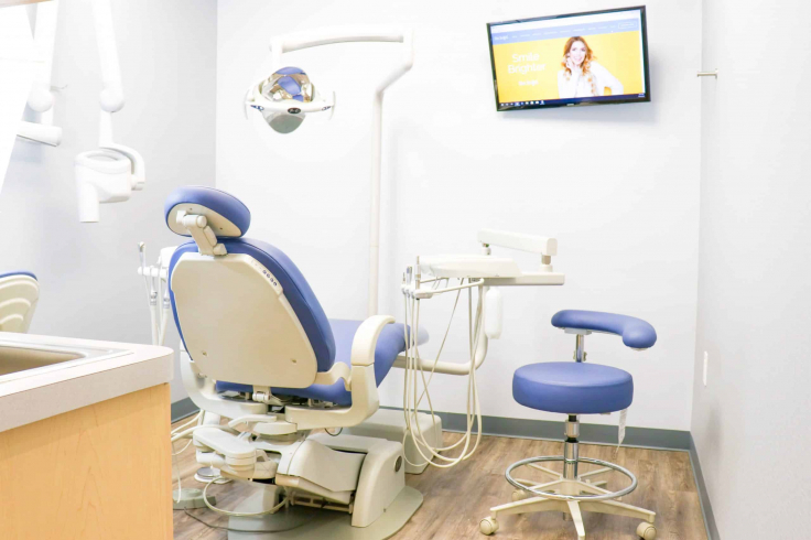 A modern dental examination room featuring blue dental chairs, overhead lighting, and a screen displaying a dental-related website.