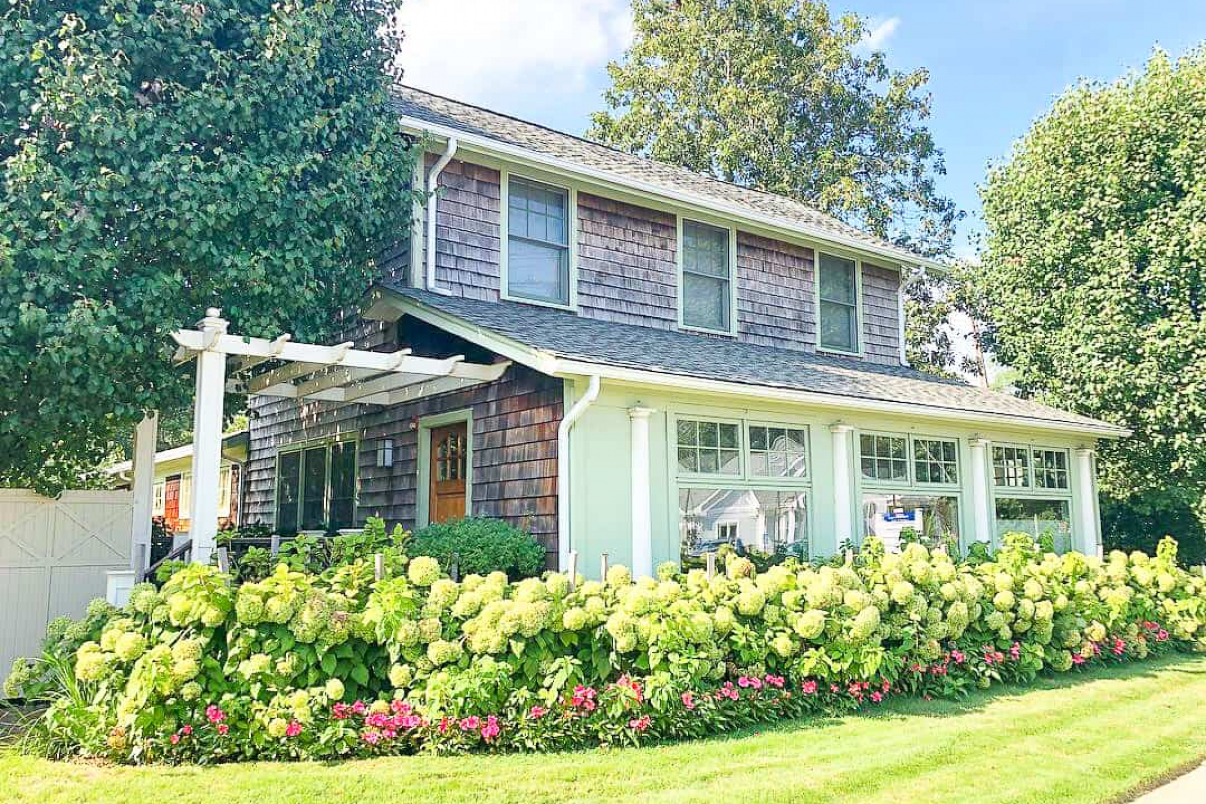 A charming two-story house with wooden shingles, surrounded by lush greenery and colorful flowers.