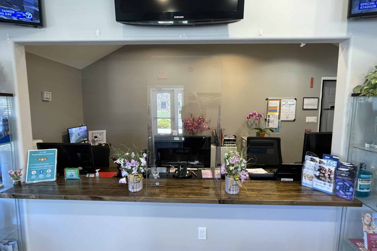 Front desk of a dental clinic featuring a clear acrylic divider, computer monitors, and decorative floral arrangements.