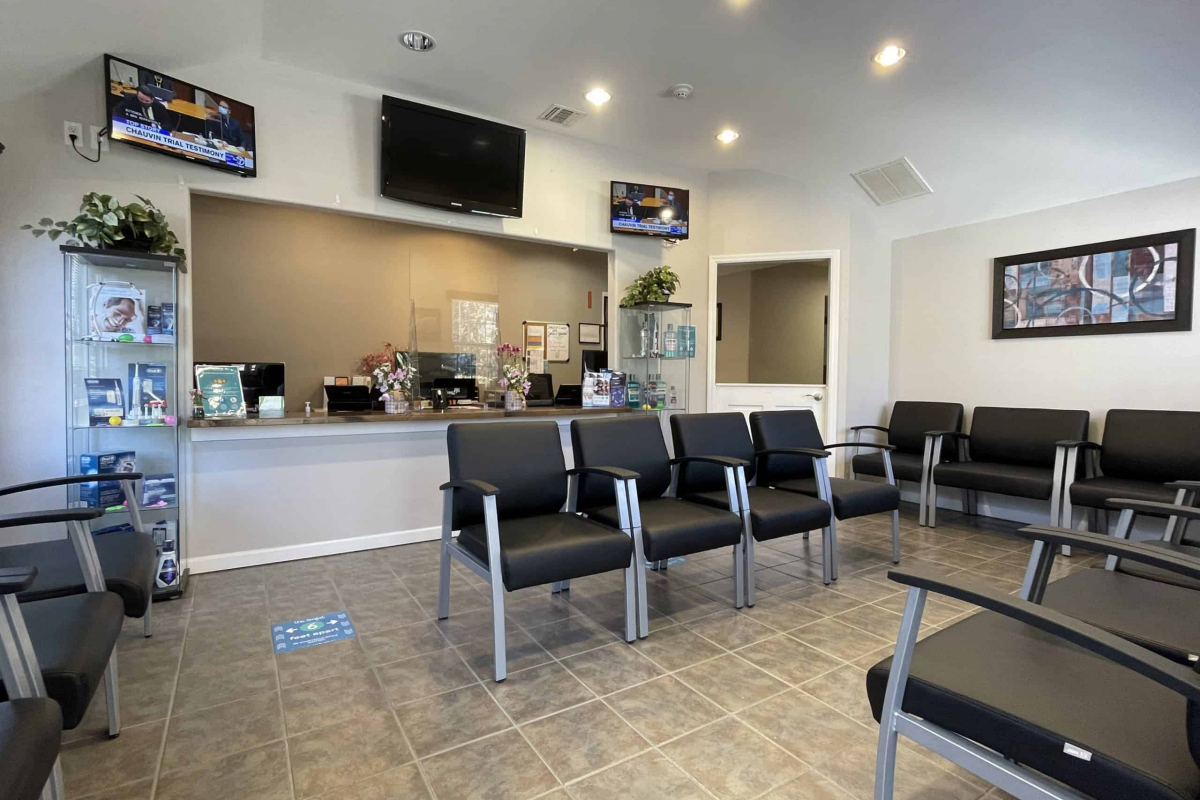 A modern dental clinic waiting area with black chairs, a reception desk with floral decorations, and multiple televisions displaying news broadcasts.