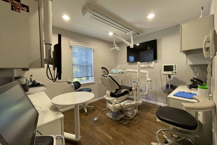 The image shows a modern dental treatment room equipped with an examination chair, dental tools, a monitor, and cabinets.