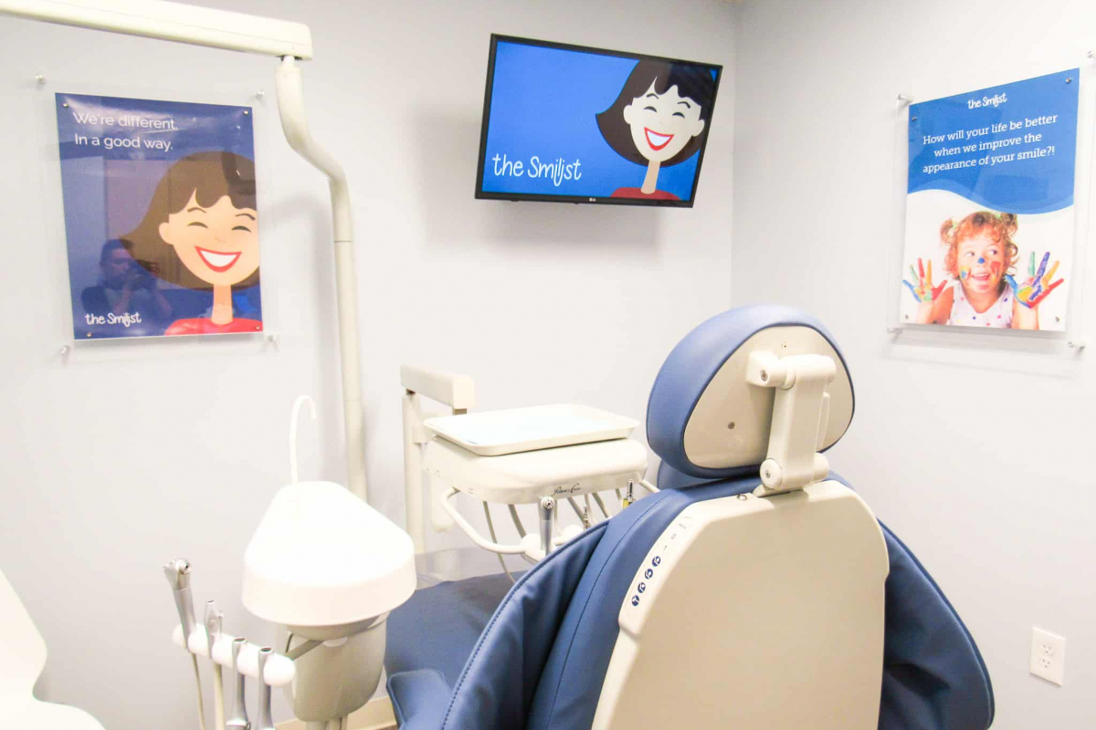A modern dental clinic room with a dental chair, equipment, and promotional posters for The Smilist on the walls.