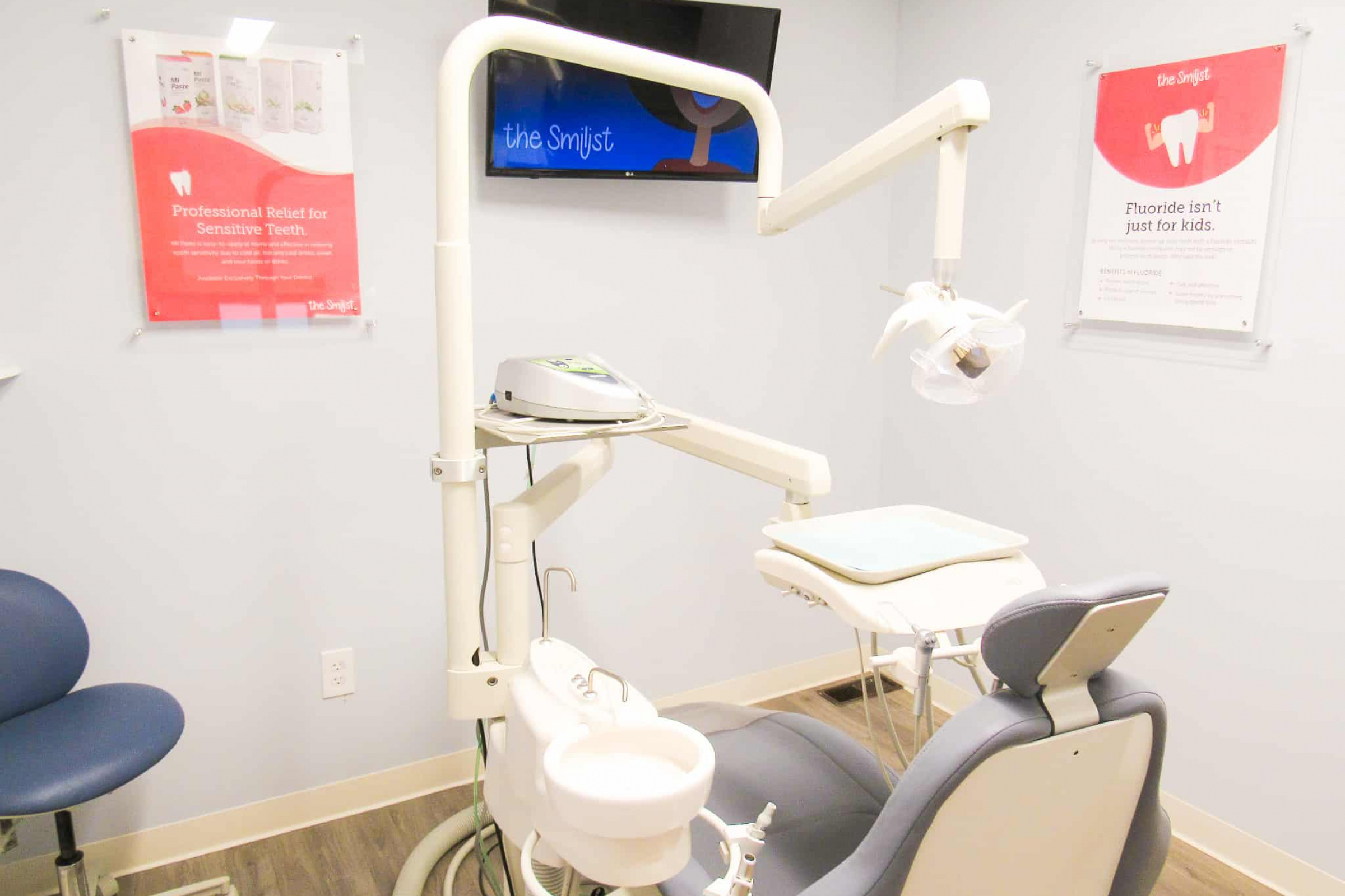 A modern dental examination room featuring a dental chair, dental equipment, and informational posters on the wall with "the Smilist" logo.
