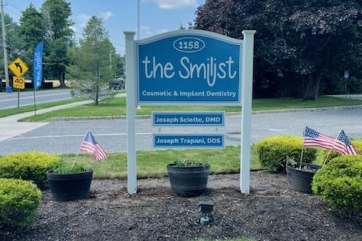 A sign for the Smilist Cosmetic & Implant Dentistry displaying the names Joseph Sciotto, DMD, and Joseph Trapani, DDS, surrounded by landscaping and American flags.