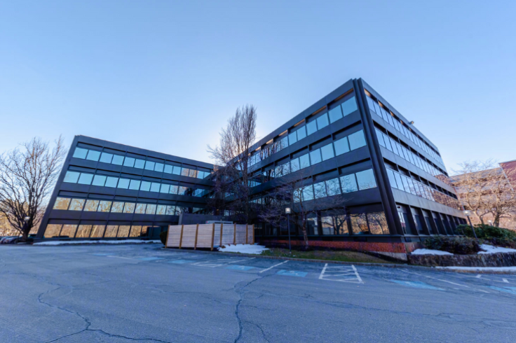 The exterior of a modern, glass-paneled office building surrounded by a parking lot and trees under a clear blue sky.